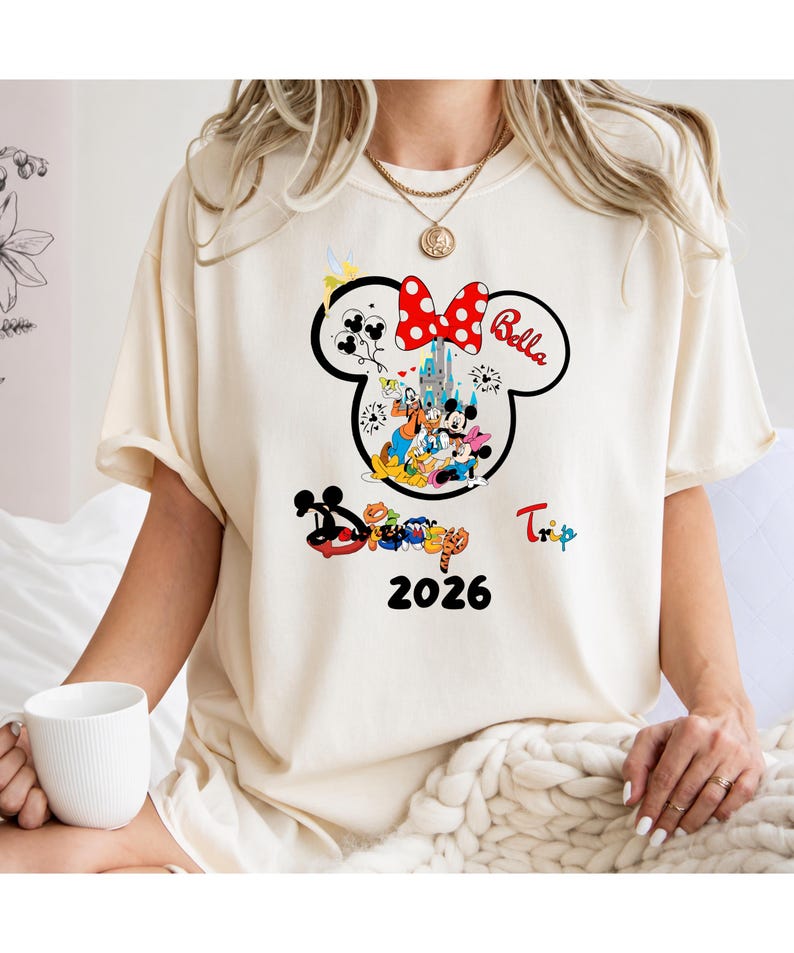 K&ouml;nnte beinhalten: Cremefarbenes T-Shirt mit einem Minnie-Mouse-Kopf-Design mit roter Schleife mit wei&szlig;en Punkten. Das Design zeigt Disney-Figuren, den Namen "Bella", das Wort "Trip" und das Jahr "2026".