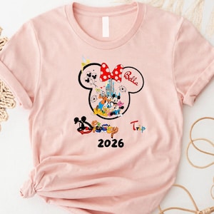 K&ouml;nnte beinhalten: Hellrosa T-Shirt mit Minnie Mouse-Design, roter Polka-Dot-Schleife und dem Namen "Bella". Das Design zeigt Disney-Figuren, ein Schloss und das Jahr "2026". Das Wort "Trip" ist ebenfalls sichtbar.