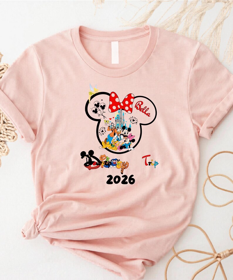 K&ouml;nnte beinhalten: Hellrosa T-Shirt mit Minnie Mouse-Design, roter Polka-Dot-Schleife und dem Namen "Bella". Das Design zeigt Disney-Figuren, ein Schloss und das Jahr "2026". Das Wort "Trip" ist ebenfalls sichtbar.