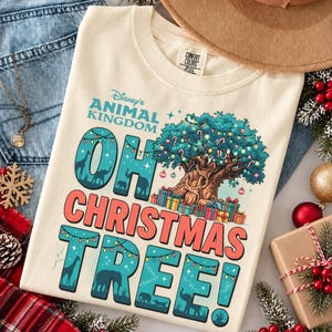 May include: A cream-coloured t-shirt featuring a graphic of the Disney Animal Kingdom Tree of Life decorated with Christmas lights and ornaments, surrounded by gifts. The text reads Disney's Animal Kingdom OH CHRISTMAS TREE!