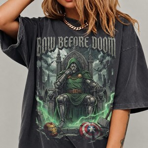 Może przedstawiać: Ciemnoszary t-shirt oversize z nadrukiem graficznym przedstawiającym zamaskowaną postać w zielonej zbroi siedzącą na tronie. Powyżej znajduje się napis BOW BEFORE DOOM. Poniżej widoczny jest czerwono-złoty hełm oraz okrągła tarcza w trzech kolorach.