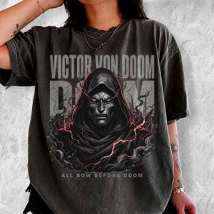 May include: A charcoal grey short-sleeve t-shirt featuring a graphic of a masked figure in a hooded cloak surrounded by red lightning and smoke. The text VICTOR VON DOOM is printed above, and the phrase ALL BOW BEFORE DOOM is printed below.