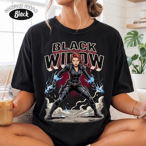 May include: A black t-shirt featuring a graphic print of the character Black Widow in a tactical suit with blue electrical energy effects. The text "BLACK WIDOW" is printed in large, bold letters above the character. A "COMFORT COLOR Black" label is visible.