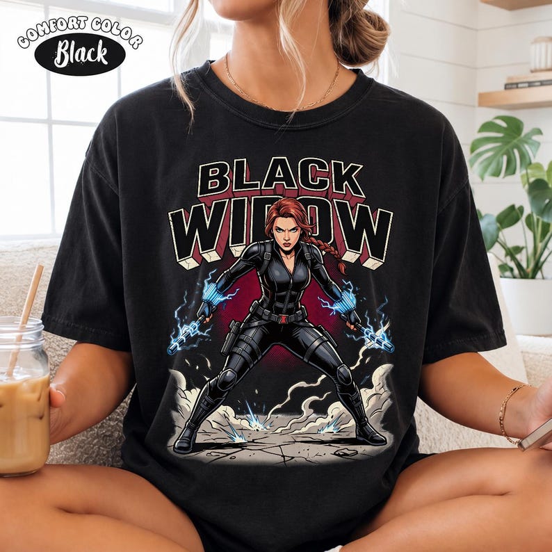 May include: A black t-shirt featuring a graphic print of the character Black Widow in a tactical suit with blue electrical energy effects. The text "BLACK WIDOW" is printed in large, bold letters above the character. A "COMFORT COLOR Black" label is visible.