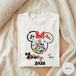 K&ouml;nnte beinhalten: Wei&szlig;es T-Shirt mit einem Minnie-Mouse-Design, einer roten Schleife mit Punkten und dem Namen "Bella". Das Design zeigt Disney-Figuren und ein Schloss, mit dem Wort "Trip" und dem Jahr "2026".