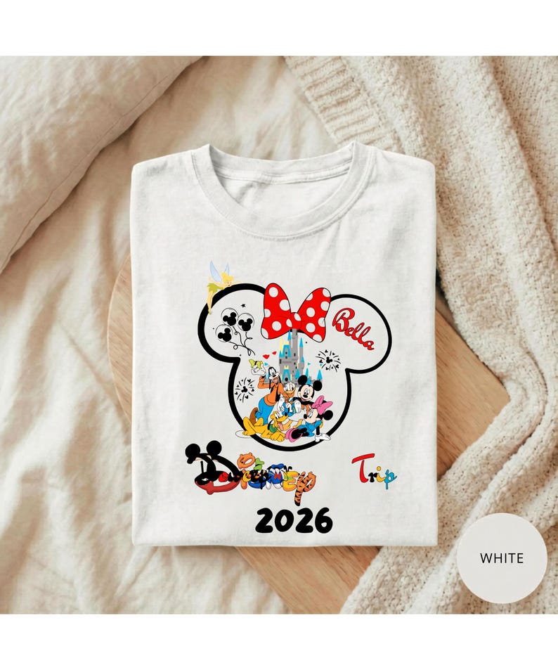 K&ouml;nnte beinhalten: Wei&szlig;es T-Shirt mit einem Minnie-Mouse-Design, einer roten Schleife mit Punkten und dem Namen "Bella". Das Design zeigt Disney-Figuren und ein Schloss, mit dem Wort "Trip" und dem Jahr "2026".