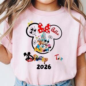 K&ouml;nnte beinhalten: Hellrosa T-Shirt mit einem Minnie Mouse-Design, einschlie&szlig;lich einer roten Schleife mit Punkten und dem Namen "Bella". Das Design zeigt Disney-Figuren, ein Schloss und das Jahr 2026. Das Wort "Trip" ist ebenfalls sichtbar.