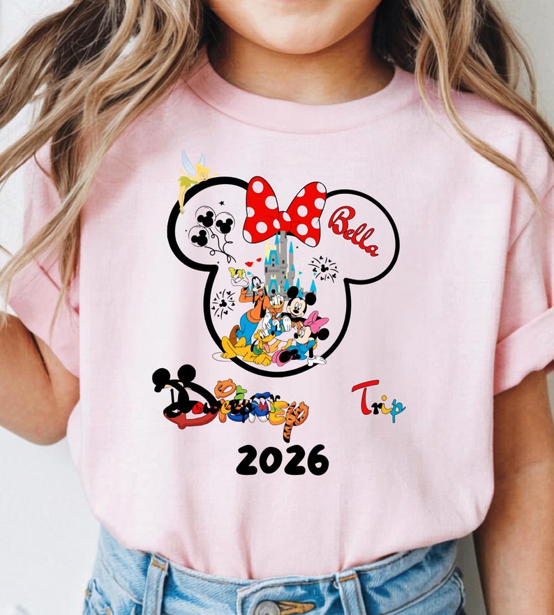 K&ouml;nnte beinhalten: Hellrosa T-Shirt mit einem Minnie Mouse-Design, einschlie&szlig;lich einer roten Schleife mit Punkten und dem Namen "Bella". Das Design zeigt Disney-Figuren, ein Schloss und das Jahr 2026. Das Wort "Trip" ist ebenfalls sichtbar.