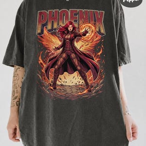 May include: A charcoal grey short-sleeve t-shirt featuring a graphic of a red-haired character in a long coat surrounded by flames. The word PHOENIX is printed in large letters above the character. A circular logo reads COMFORT COLORS Pepper.
