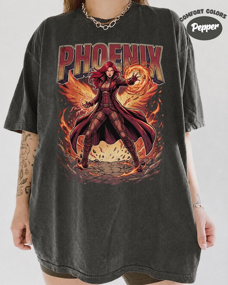 May include: A charcoal grey short-sleeve t-shirt featuring a graphic of a red-haired character in a long coat surrounded by flames. The word PHOENIX is printed in large letters above the character. A circular logo reads COMFORT COLORS Pepper.