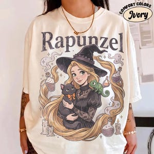 Könnte beinhalten: Ein elfenbeinfarbenes T-Shirt mit einer Grafik einer Figur mit langem blonden Haar und Hexenhut, die eine schwarze Katze mit orangefarbener Schleife und eine grüne Eidechse auf der Schulter hält. Darüber steht der Text Rapunzel mit Zaubertrankflaschen.