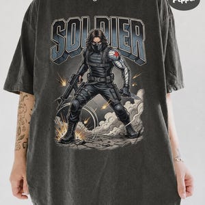 May include: A charcoal grey t-shirt featuring a graphic of a masked soldier with a metallic arm and a red star emblem. The soldier holds a rifle and a knife amidst smoke and sparks. The word SOLDIER is printed in large block letters above the figure.