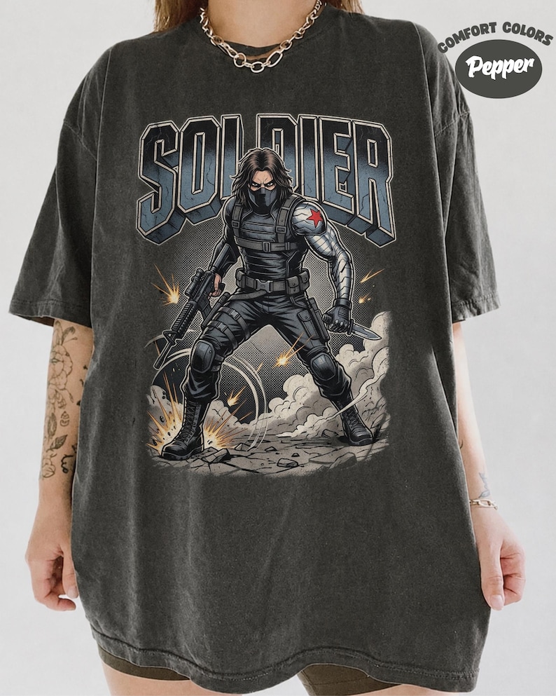 May include: A charcoal grey t-shirt featuring a graphic of a masked soldier with a metallic arm and a red star emblem. The soldier holds a rifle and a knife amidst smoke and sparks. The word SOLDIER is printed in large block letters above the figure.