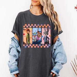 Może przedstawiać: Ciemnoszara koszulka z kolorową grafiką postaci z filmu Zwierzogr&oacute;d. Grafika jest obramowana wzorem w kratkę w odcieniach r&oacute;żu, pomarańczy i fioletu. Koszulka ma swobodny styl.