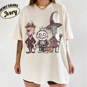 May include: An ivory short-sleeve t-shirt featuring a graphic print of three cartoon characters in a sketch style. The characters include a pirate, a crowned figure with a lollipop, and a witch holding a broom. Text reads COMFORT COLORS IVORY.