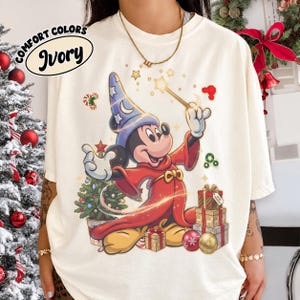 Könnte beinhalten: Ein elfenbeinfarbenes T-Shirt mit einer Grafik einer Zeichentrickmaus in blauem Zaubererhut und rotem Gewand, die einen Zauberstab hält. Das Design zeigt einen Weihnachtsbaum, gestapelte Geschenke und goldene Sterne. Text lautet COMFORT COLORS IVORY.