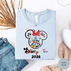 K&ouml;nnte beinhalten: Hellblaues T-Shirt mit einem Minnie Mouse-Design, einer roten Schleife mit Punkten und dem Namen "Bella". Das Design enth&auml;lt ein Schloss, Figuren und das Jahr "2026". Das Wort "Trip" ist ebenfalls sichtbar. Das T-Shirt ist gefaltet.