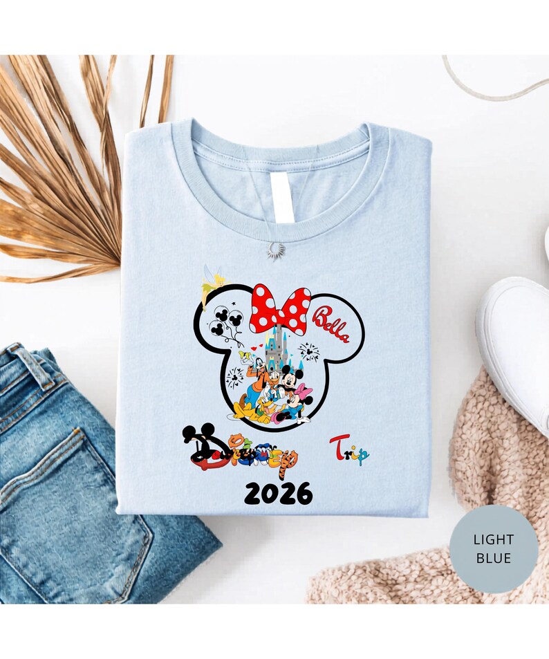 K&ouml;nnte beinhalten: Hellblaues T-Shirt mit einem Minnie Mouse-Design, einer roten Schleife mit Punkten und dem Namen "Bella". Das Design enth&auml;lt ein Schloss, Figuren und das Jahr "2026". Das Wort "Trip" ist ebenfalls sichtbar. Das T-Shirt ist gefaltet.