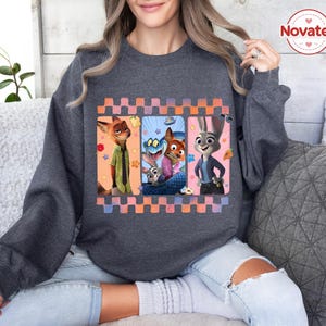 Może przedstawiać: Ciemnoszara bluza z kolorową grafiką postaci z filmu animowanego Zwierzogr&oacute;d. Grafika zawiera Nicka Wilde'a, Judy Hopps i inne postacie w obramowanej kompozycji. Bluza ma długie rękawy i okrągły dekolt.