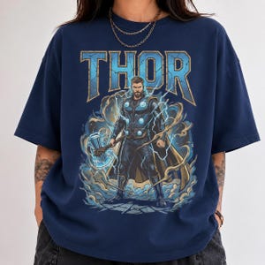 May include: A navy blue short-sleeve t-shirt featuring a graphic print of the character Thor. The design includes the text THOR in large blue letters above the character, who is holding a hammer surrounded by blue lightning and swirling clouds.