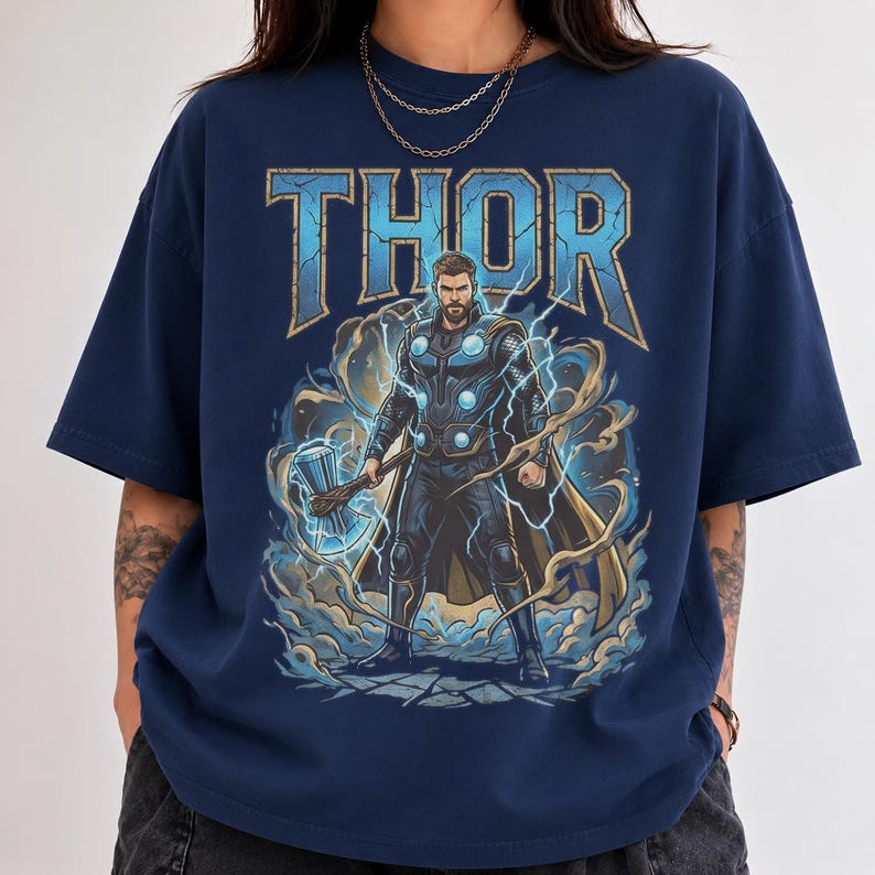 May include: A navy blue short-sleeve t-shirt featuring a graphic print of the character Thor. The design includes the text THOR in large blue letters above the character, who is holding a hammer surrounded by blue lightning and swirling clouds.