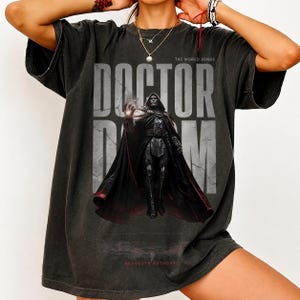 May include: A charcoal grey short-sleeve t-shirt featuring a large graphic print of the character Doctor Doom. The design includes the text DOCTOR DOOM in large letters, the phrase THE WORLD BENDS, and the text ABSOLUTE AUTHORITY at the bottom.