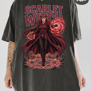 May include: A charcoal grey t-shirt featuring a graphic of the Scarlet Witch in a red costume with a flowing cape, surrounded by red magical energy and a glowing circular rune on the ground. The text "SCARLET WITCH" is printed above in red.