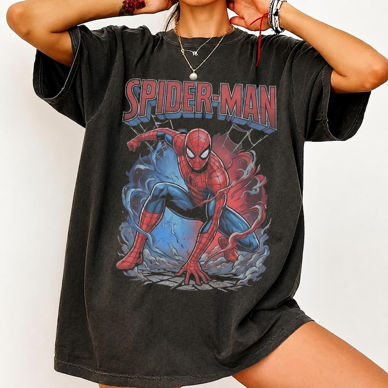 May include: A charcoal grey short-sleeve t-shirt featuring a graphic print of the Marvel character Spider-Man in a crouching pose. The text SPIDER-MAN is printed in bold red letters above the character, surrounded by blue and red smoke effects.