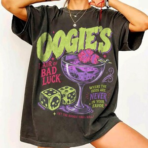 Könnte beinhalten: Ein anthrazitfarbenes Kurzarm-T-Shirt mit einem Grafikdruck, der den Text OOGIE'S LAIR OF BAD LUCK, WHERE THE ODDS ARE NEVER IN YOUR FAVOR und LET THE BOOGIE TIMES ROLL sowie Illustrationen von Würfeln und einem Cocktailglas mit Totenkopf zeigt.