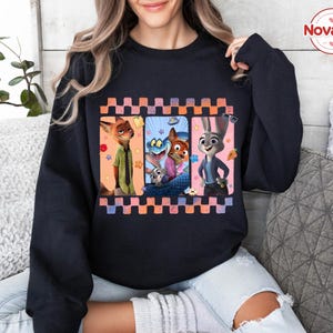 Może przedstawiać: Czarna bluza z kolorową grafiką postaci z filmu animowanego Zwierzogr&oacute;d. Projekt zawiera Nicka Wilde'a, Judy Hopps i inne postacie w oprawie. Słowo "Novatee" znajduje się w okręgu w prawym g&oacute;rnym rogu.