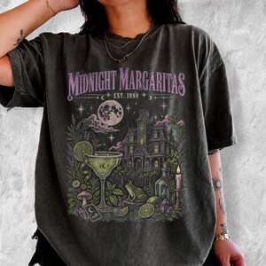 Puede incluir: Una camiseta gris carbón con un estampado gráfico que incluye el texto "MIDNIGHT MARGARITAS EST. 1998". El diseño muestra una gran casa encantada, una luna llena, una copa de margarita con una rodaja de lima, una rana, una vela y varias plantas.