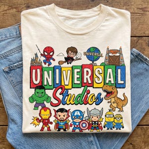 May include: A cream-coloured t-shirt featuring a colourful graphic print of Universal Studios characters and icons. The design includes Spider-Man, Harry Potter, the Hulk, Iron Man, Doctor Strange, Captain America, Thor, Minions, and a T-Rex.