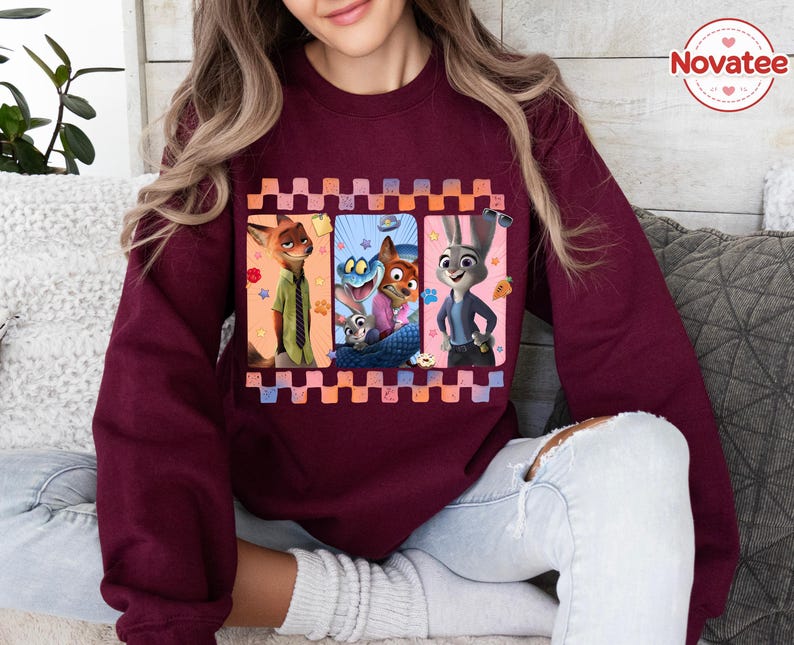 Może przedstawiać: Bordowa bluza z kolorową grafiką postaci z animowanego filmu Zwierzogr&oacute;d. Grafika podzielona jest na trzy panele z postaciami Nicka Wilde'a, Judy Hopps i innymi. Bluza ma okrągły dekolt i długie rękawy.