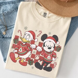 May include: A cream-coloured t-shirt featuring a graphic of Donald Duck and Mickey Mouse wearing Santa hats and Christmas jumpers. The design includes red and white candy canes, green gift boxes, and snowflakes over a red checkered background.