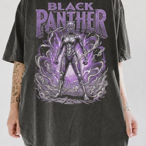 May include: A charcoal grey oversized t-shirt featuring a graphic print of the Black Panther character in a purple and silver suit. The text "BLACK PANTHER" is printed in large purple letters above the character, surrounded by purple energy swirls.