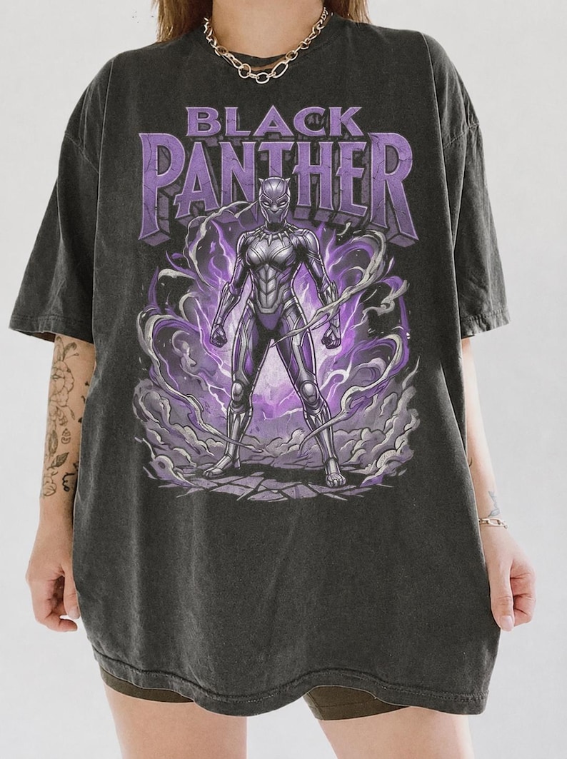 May include: A charcoal grey oversized t-shirt featuring a graphic print of the Black Panther character in a purple and silver suit. The text "BLACK PANTHER" is printed in large purple letters above the character, surrounded by purple energy swirls.