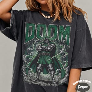 May include: A charcoal grey short-sleeve t-shirt featuring a graphic print of the character Doctor Doom in a green cape and metallic armour. The word DOOM is printed in large green block letters above the character, surrounded by swirling smoke.