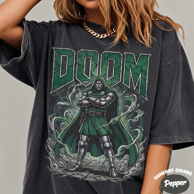 May include: A charcoal grey short-sleeve t-shirt featuring a graphic print of the character Doctor Doom in a green cape and metallic armour. The word DOOM is printed in large green block letters above the character, surrounded by swirling smoke.