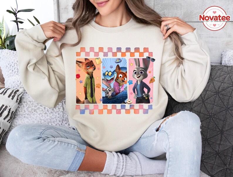 Może przedstawiać: Beżowa bluza z kolorową grafiką postaci z animowanego filmu Zwierzogr&oacute;d. Grafika jest obramowana wzorem w kratkę w odcieniach r&oacute;żu, pomarańczy i błękitu. Bluza w stylu wygodnym i swobodnym.