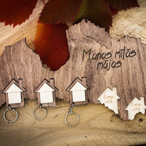 May include: A wooden key holder shaped like the outline of Latvia. It features three small houses with key rings and the text "Manas mīlās mājas" which translates to "My beloved home" in English. The key holder is on a wooden surface with fall leaves.