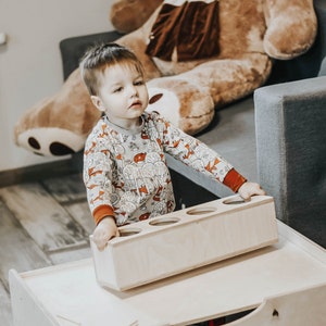 May include: A young child in a beige and orange patterned long-sleeved shirt is playing with a wooden toy with four round holes. The toy is sitting on a wooden table with a drawer.
