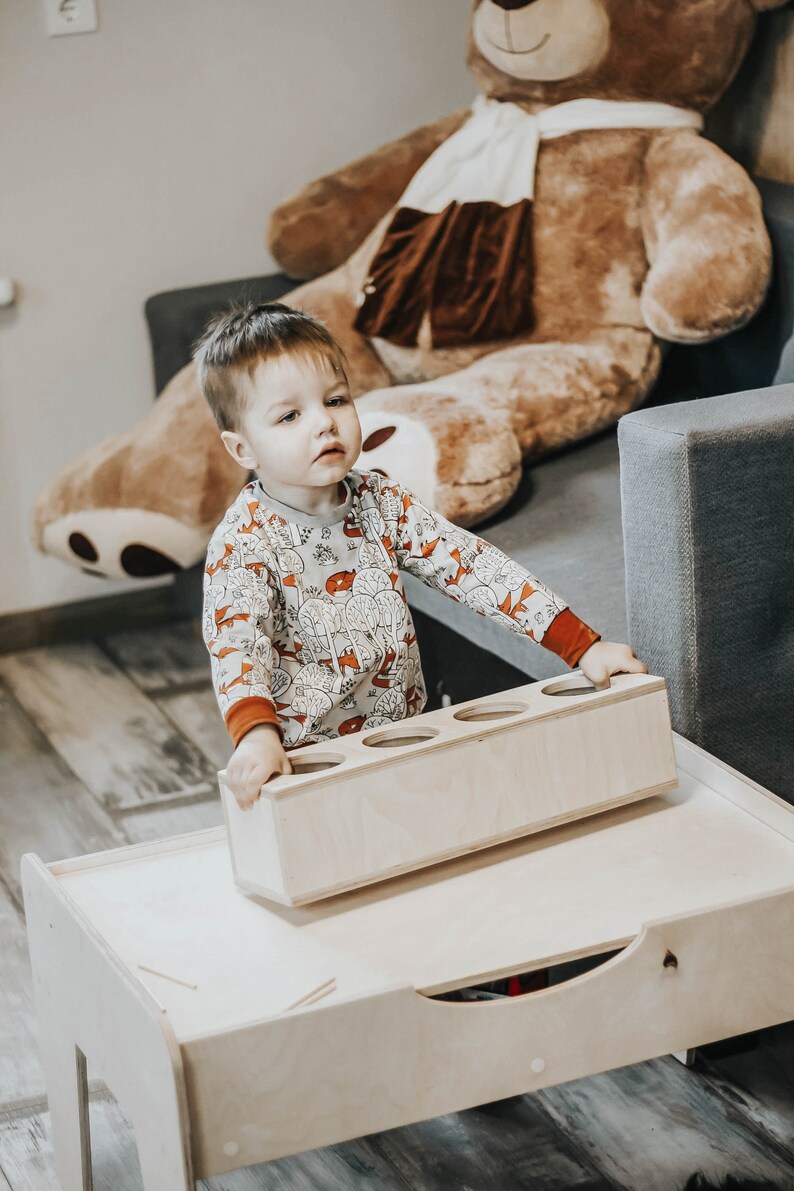 May include: A young child in a beige and orange patterned long-sleeved shirt is playing with a wooden toy with four round holes. The toy is sitting on a wooden table with a drawer.