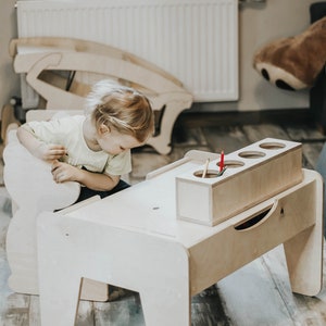 May include: A child sits at a wooden table with a built-in storage compartment for crayons and pencils. The table is light brown and has a simple design.