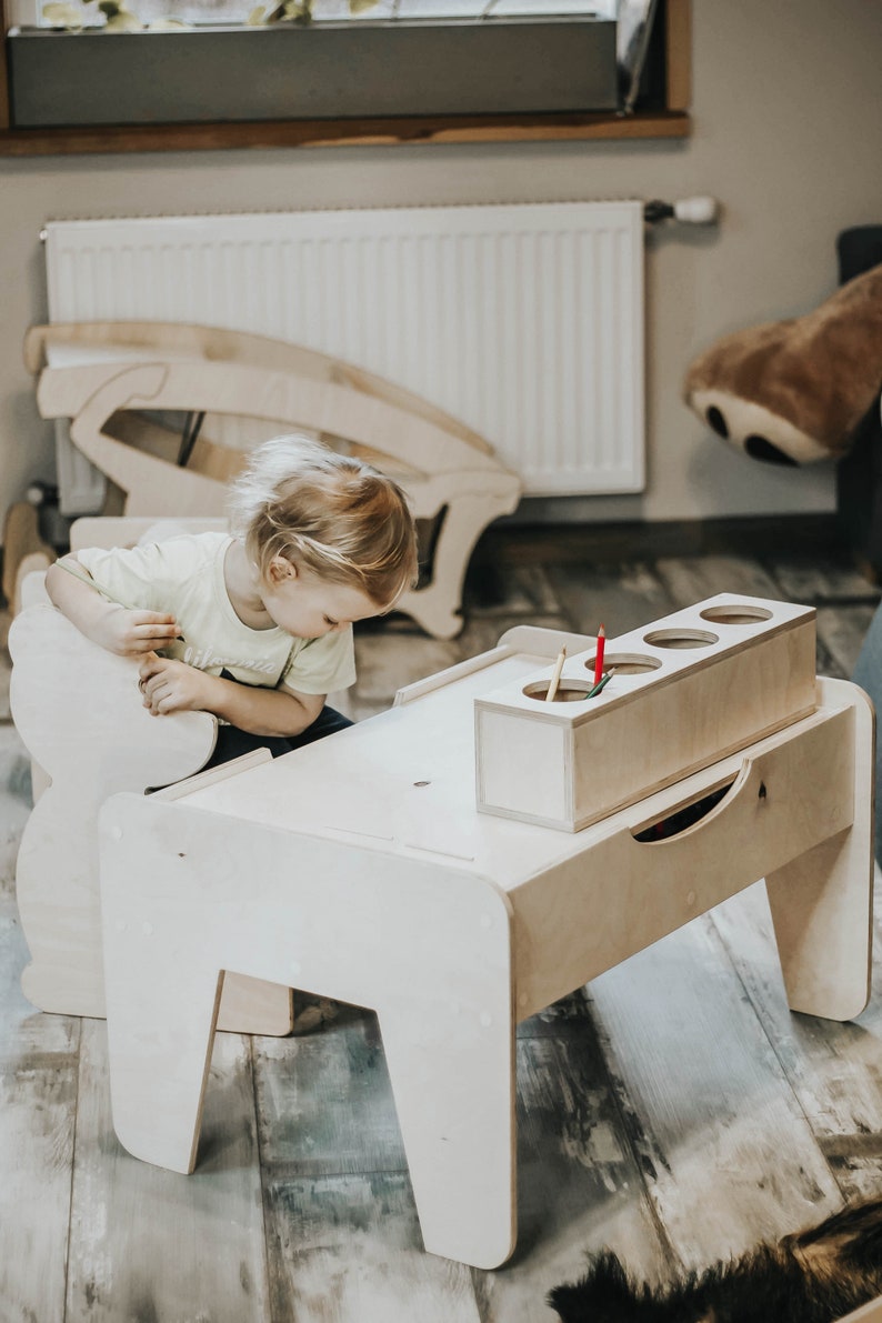 May include: A child sits at a wooden table with a built-in storage compartment for crayons and pencils. The table is light brown and has a simple design.