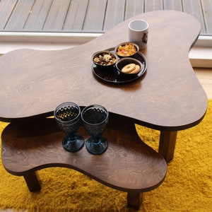 Puede incluir: Dos mesas de anidaci&oacute;n de madera con una forma irregular y org&aacute;nica. La mesa superior tiene una taza blanca, dos cuencos peque&ntilde;os con aperitivos y dos vasos de vidrio azul. La mesa inferior es marr&oacute;n y tiene una alfombra peluda amarilla debajo.