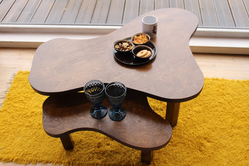 Puede incluir: Dos mesas de anidaci&oacute;n de madera con una forma irregular y org&aacute;nica. La mesa superior tiene una taza blanca, dos cuencos peque&ntilde;os con aperitivos y dos vasos de vidrio azul. La mesa inferior es marr&oacute;n y tiene una alfombra peluda amarilla debajo.