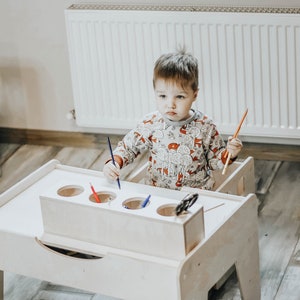 May include: A white wooden children's table and chair set with a built-in pencil holder. The table has a drawer and four round holes for holding pencils. A young child is sitting at the table, holding a pencil and looking at the camera.