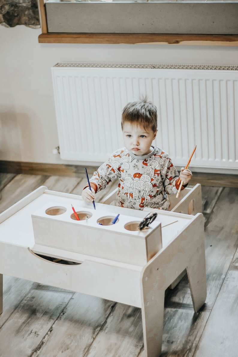 May include: A white wooden children's table and chair set with a built-in pencil holder. The table has a drawer and four round holes for holding pencils. A young child is sitting at the table, holding a pencil and looking at the camera.
