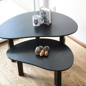 May include: Two black nesting coffee tables with a unique organic shape. The top table has a white mug with a wolf design and a book on top. The bottom table has a group of pine cones on top.