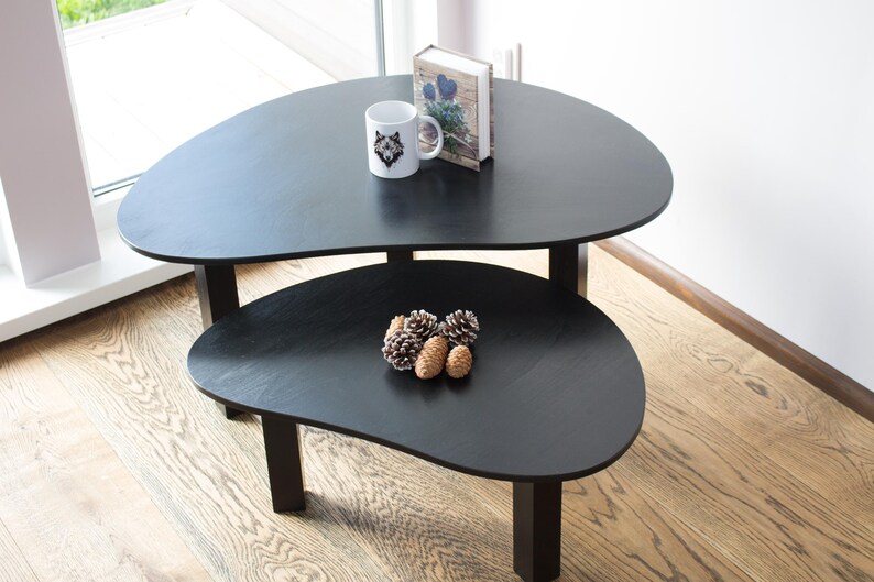 May include: Two black nesting coffee tables with a unique organic shape. The top table has a white mug with a wolf design and a book on top. The bottom table has a group of pine cones on top.